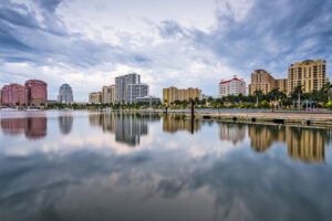 Skyline of West Palm Beach, Florida at dusk, featuring modern high-rise buildings, palm trees, and their reflections on the calm waterfront under a cloudy sky.